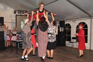 Gruppe von Tanzern hebt einen Mann hoch, alle in farbenfroher Kleidung.