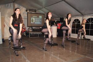 Frauen in schwarzen Kostümen tanzen auf Hockern. Sie tragen graue Strumpfhosen und schwarze Oberteile.