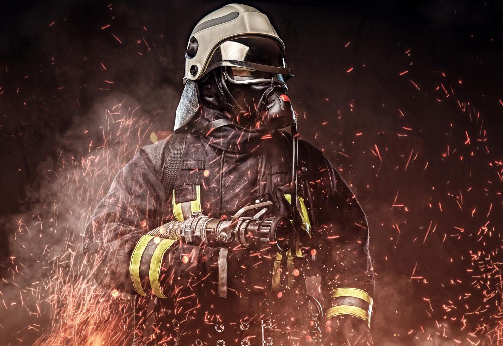 Ein Feuerwehrmann in Schutzkleidung und Helm, umgeben von Rauch und Flammen. Er atmet durch eine Maske.
