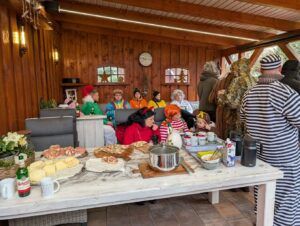 Kinder in bunten Kostümen sitzen am Buffet. Ein Erwachsener im schwarz-weißen Streifenanzug steht daneben.